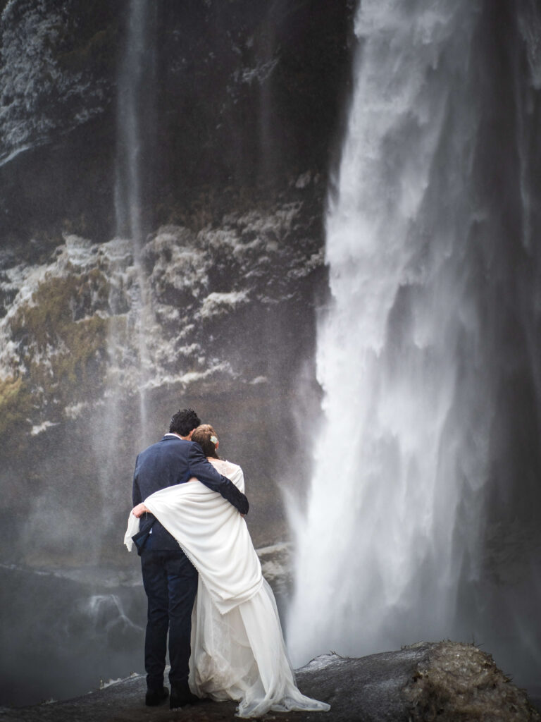 bride and groom cuddling in front of seljalandsfoss waerfall in inceland during the winter